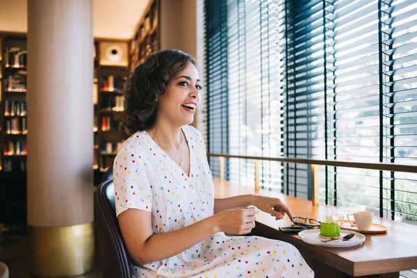 Young content lady sitting at table with delicious mousse cake and hot drink against window while looking away in cafe of book store