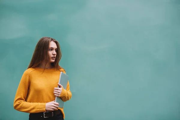 Young calm woman in casual pullover and jeans with long hair looking away standing on turquoise background while holding modern laptop