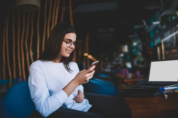 Glad female freelancer in casual outfit with toothy smile sitting in comfortable armchair and using smartphone for checking social networks during break