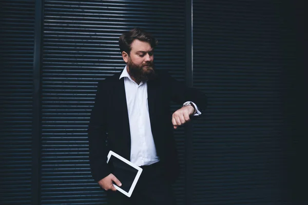 Serious bearded male in black business suit with tablet looking at time on wristwatch checking time while standing near black wall