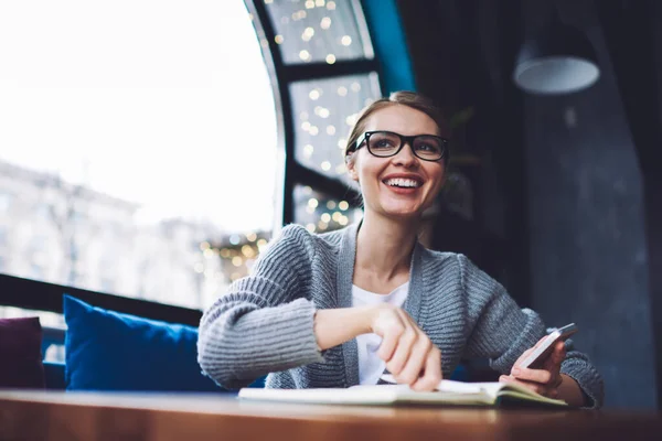 Low angle of happy woman in casual wear and glasses taking notes while browsing smartphone and looking away in cozy cafe at daytime