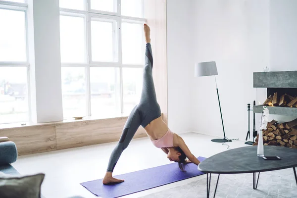 Full body unrecognizable barefoot slim lady in tight leggings doing downward yoga pose variation with one leg up while practicing home in daytime