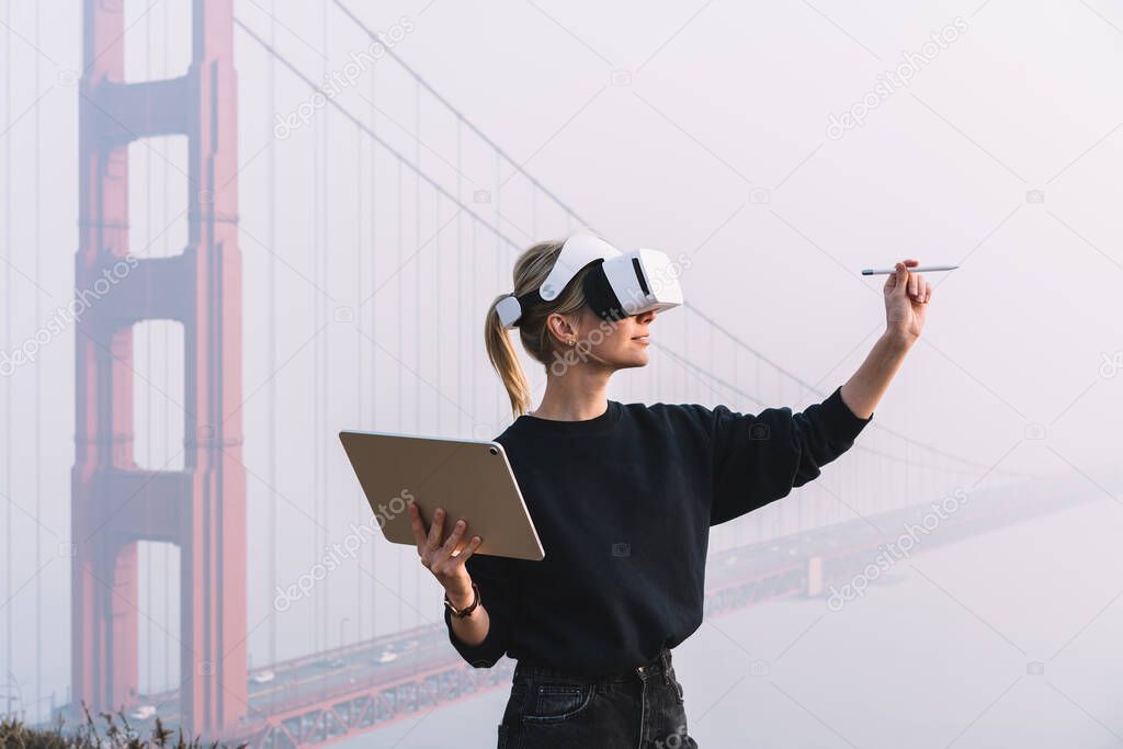 Side view of positive female designer standing on elevation in creative headset with tablet while using virtual display to draw sketch