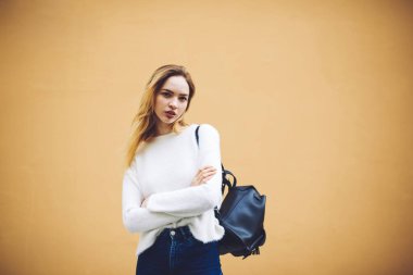 Kendine güvenen genç bir bayan, büyük kahverengi duvarın yanında dikilirken kameraya bakıyor ve beyaz kazak ve sırt çantalı kot pantolon giyiyor ve elleri çapraz.