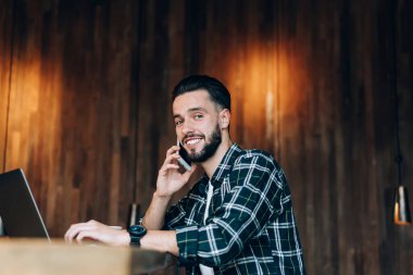 Akıllı telefon uygulaması üzerinden ararken kameraya gülümseyen dizüstü bilgisayarlı neşeli erkek metin yazarı