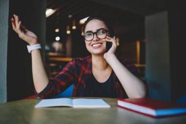 Günlük kıyafet ve gözlüklerle gülümseyen etnik kadın tele-işçi telefon kullanarak arama yapmak ve not almak için cep telefonu kullanıyor.