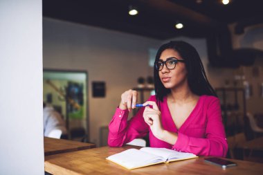Konsantre olmuş renkli günlük kıyafetli bayan öğrenci açık bir defterle masada oturmuş düşünürken gözlerini kaçırıyor.