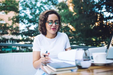 Günlük giysiler ve gözlüklü genç bir kadın yönetici, açık kafede çalışırken bir fincan kahve ve kitap okuyarak masada oturuyor.
