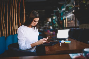 Modern çalışma alanında proje için bilgi toplarken bilgisayarını açıp akıllı telefon kullanan düşünceli serbest çalışan kadın.