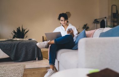 Kot pantolon ve beyaz gömlek giyen güzel bir bayan rahat koltukta bacak bacak üstüne atmış bir şekilde otururken şık bir yatak odasında dizüstü bilgisayar ve cep telefonu kullanıyor.