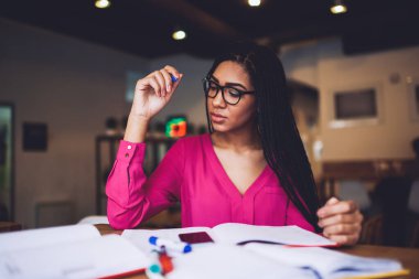 Konsantre olmuş, gözlüklü, uzun siyah saçlı Afro-Amerikan bir kız öğrenci masada oturup ödev yaparken aşağıya bakıyor.