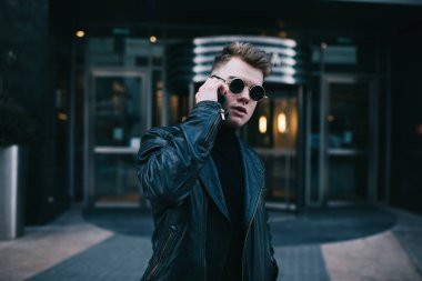 Siyah elbiseli, ciddi bir genç adam binanın girişinde duruyor ve arkadaşını beklerken telefonla konuşuyor.