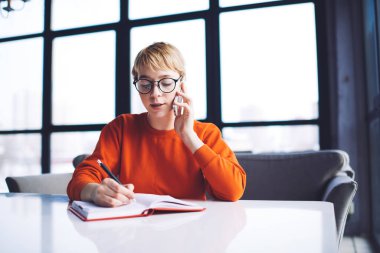 Gündelik giysiler içinde genç, konsantre bir kadın işçi. Not defterine not alıyor ve açık modern çalışma alanında çalışırken telefonla konuşuyor.