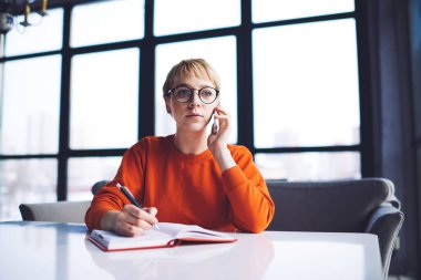 Uzaktan kumandalı, kısa saçlı, gözlüklü genç bir kadın. Masaya oturuyor, not alıyor ve cep telefonuyla konuşuyor.