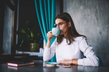 Beyaz kazaklı, gözlüklü, kahve içen, elinde not defteri ve akıllı telefonuyla kafeye bakan genç bir kadın.