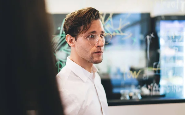 Side view of serious businessman in office wear and eyeglasses standing in modern office and looking away while listening to colleagues