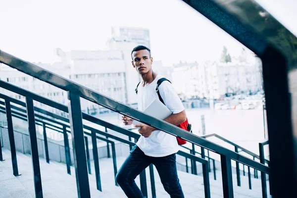 Kırmızı sırt çantalı genç bir erkek öğrencinin modern dizüstü bilgisayarı ve içecek paketini üst katta kentsel arka planda metal korkulukların yanında yürürken görüntüsü