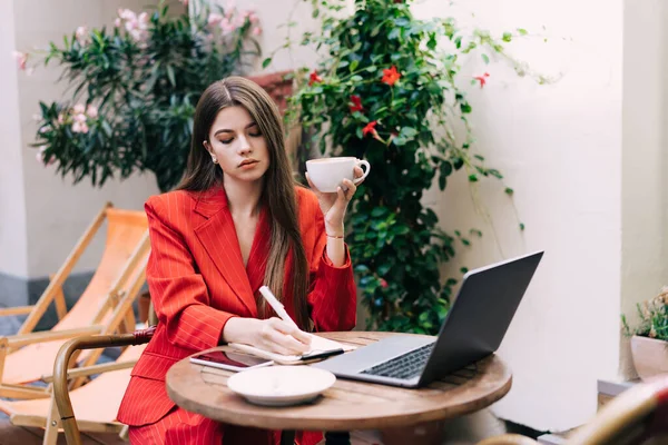 Şık giyinmiş, elinde beyaz bir fincan kahve tutan ve dizüstü bilgisayar üzerinde çalışırken deftere yazan göz kamaştırıcı bir genç bayan. 