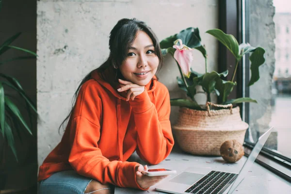 Happy Asian woman remote worker in bright hoodie looking away while sitting near window with laptop leaning on hand and using smartphone