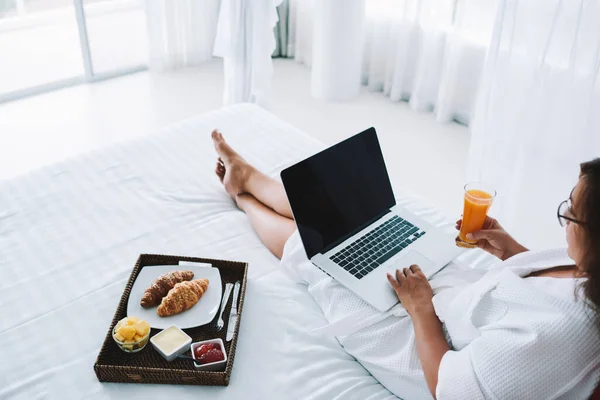 From above of crop anonymous female in white dressing gown lying on comfortable bed while using laptop and drinking juice in morning