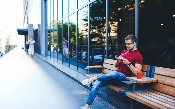 Concentrated youthful bearded guy wearing jeans sitting near modern building with takeaway drink and reading contact information on business card while messaging on smartphone