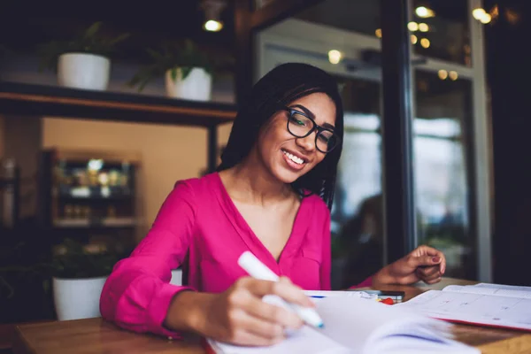 アフリカ系アメリカ人の満足度の高い女性で ノートやノートを見ながらカフェでの試験の準備をしながら カジュアルな服や眼鏡をかけています — ストック写真