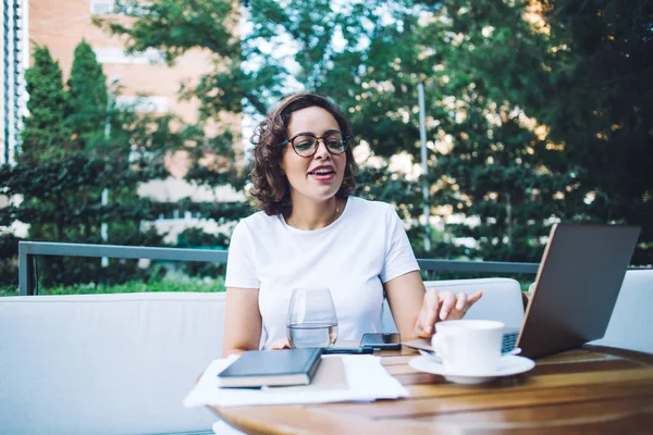 Beyaz tişörtlü, serbest çalışan mutlu genç bir bayan yazın açık hava restoranının terasında otururken netbook üzerinde çalışıyor.