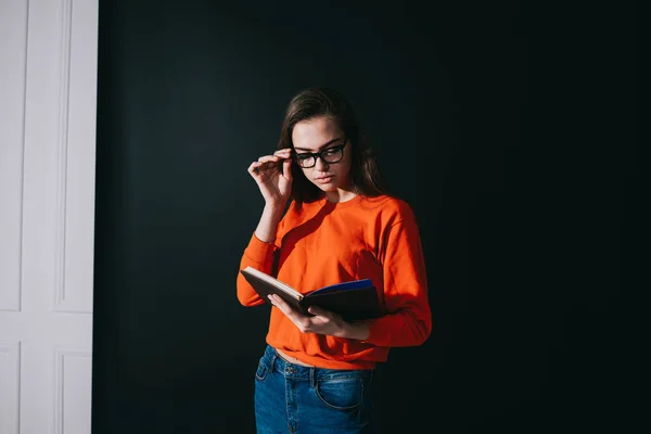 Serious dark haired female in casual sweater and jeans with eyeglasses reading notepad in black cover in studio on black background