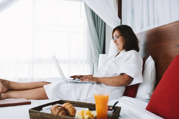 Side view of positive female in white dressing gown lying on comfortable bed and typing on laptop in hotel of Kuala Lumpur
