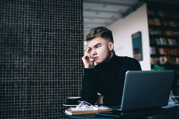 Siyah balıkçı yaka yakalı dalgın genç adam kütüphaneden uzak bir projede çalışırken kaşlarını okşuyor ve yüzünü çeviriyor.