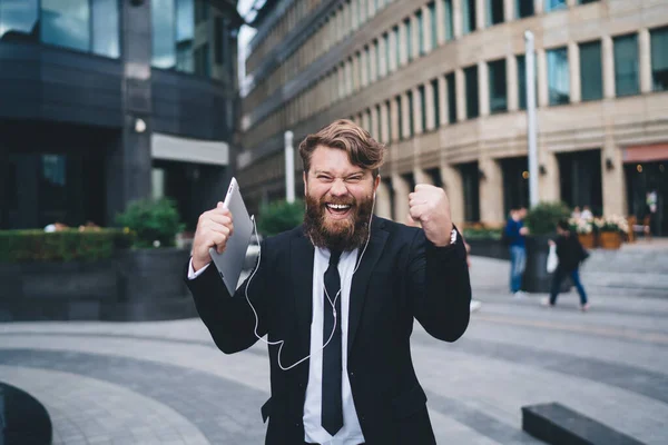 Happy bearded male in formal clothes with tablet and earphones looking at camera while standing on street near modern buildings