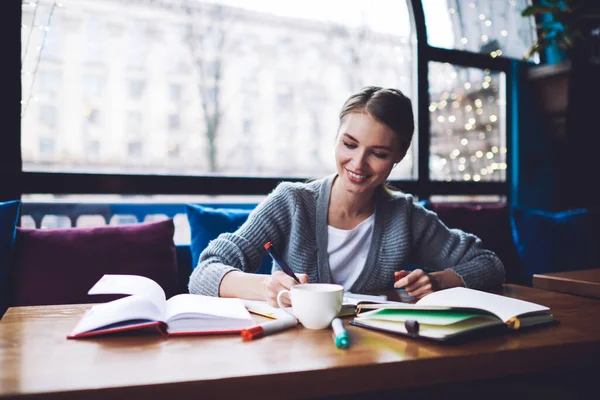 Dişli tebessümü olan mutlu bayan öğrenci ders kitaplarıyla masada otururken aşağıya bakıyor ve kalemle bilgiyi parlaklaştırıyor.