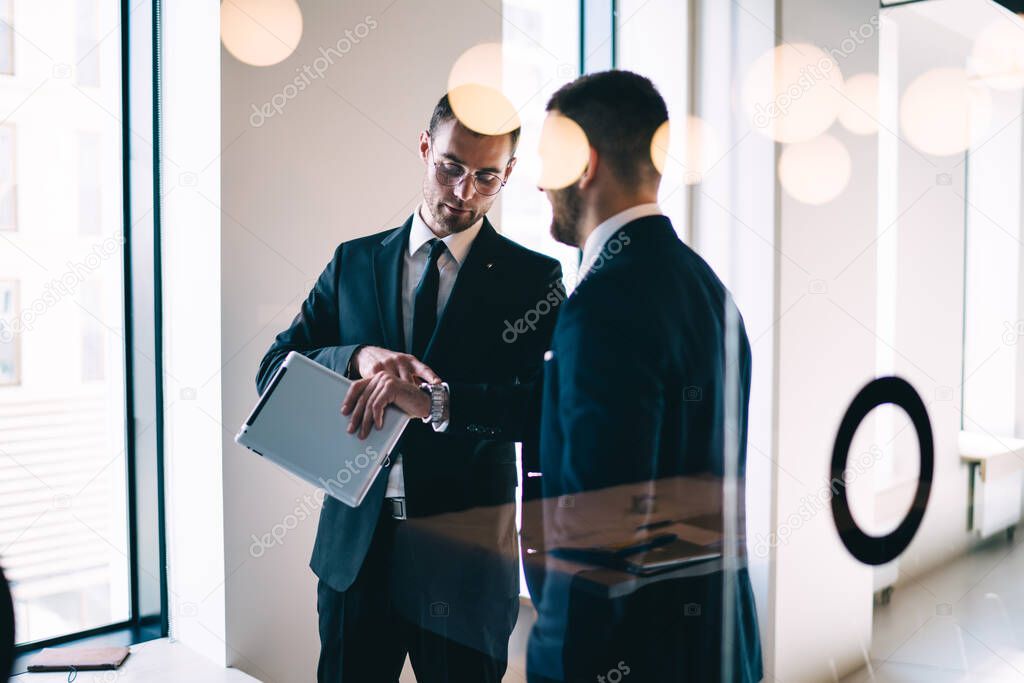 Through glass of serious young businessman with tablet in hand in formal suit checking time on wristwatch while standing in office corridor 