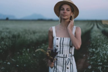Lavanta tarlasında alacakaranlıkta dikilip elinde çiçeklerle kameraya bakan genç bir kadın.