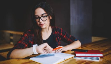 Gözlüklü konsantre kadın uzaktan çalışan Kol saatine bakıyor ve gündüzleri kafede elinde defterlerle masada oturuyor.