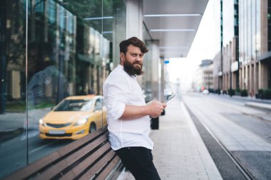 Şehir caddesinde ulaşımı beklerken modern tabletlere göz atan resmi kıyafetler giyen sakallı adamın yan görüntüsü. Ve kameraya bakıyor.