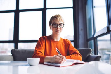 Uzaktan kumandalı kadın çalışanın pencerenin yanındaki masada oturup kahve fincanı ile kafedeki projede notlar yazmasına sevindim.