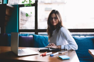 Kafeteryadaki rahat koltukta oturan gözlüklü genç bir kadın. Ahşap masa, keçeli kalem, dizüstü bilgisayar ve kameraya bakıyor.