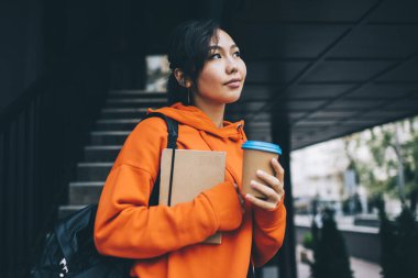 Canlı kapüşonlu Asyalı düşünceli bayan öğrenci. Sırt çantasıyla sokakta elinde not defteri ve bir fincan kahveyle dikilirken başka yere bakıyor.