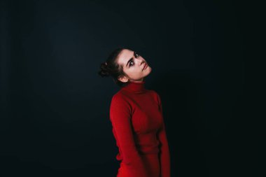 Saçları topuz şeklinde toplanmış, siyah arka planda dikilirken kameraya ve eğimli kafaya bakan genç model.