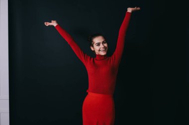 Kırmızı kazaklı ve etekli neşeli genç bir kadın gri arka planda el kaldırıyor ve kameraya bakıyor.