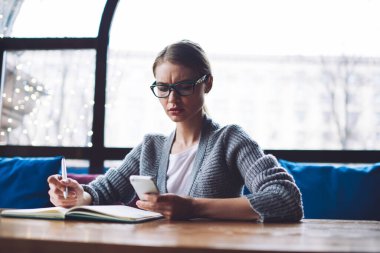 Düşünceli bayan öğrenci somurtuyor ve elinde defterle masada oturuyor. Cep telefonundan ödev yaparken bildirimleri kontrol ediyor.