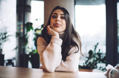 Modern kafede ahşap masada oturan ve kameraya dil gösterirken kameraya bakan günlük kıyafetli mutlu kadın.