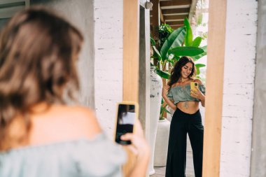 Modayı takip eden mutlu, modaya uygun bir kadın gülümseyerek ve cep telefonuyla fotoğraf çekerken aynanın yanında neşeyle dikiliyor.