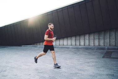 Akıllı telefon ve kulaklıklarla koşan genç sporcunun müzik dinlerken ve sabah antrenmanında gözlerini kaçırırken yan görüntüsü