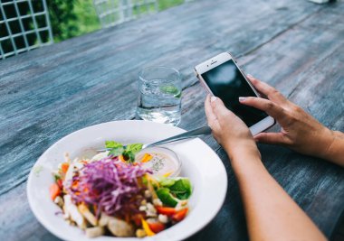 Tanımlanamayan bir kadının taze sebze salatası ve bir bardak suyla sağlıklı öğle yemeği yemesi ve akıllı telefondan mesajlaşması.