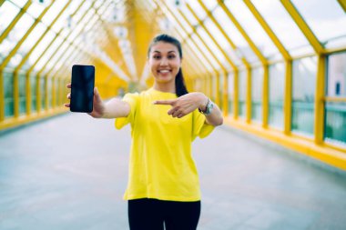 Neşeli etnik kadın, günlük kıyafetler içinde, dişlek tebessümle çatıda duruyor ve kameraya bakarken yeni bir akıllı telefonu işaret ediyor.