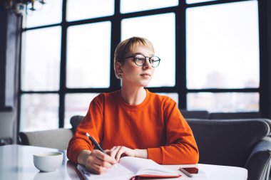 Gözlüklü, kısa saçlı, rahat giyinen, masa başında not defteriyle oturan serbest çalışan genç bir kadın.