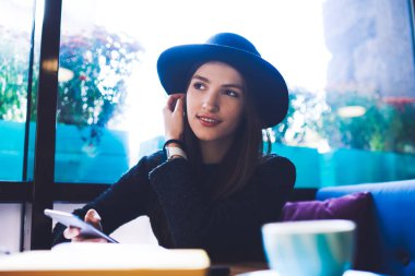 Zarif kıyafetli, siyah şapkalı, elinde kahve fincanı, kulağının arkasında saç saklayan akıllı telefonlu bir kadın.