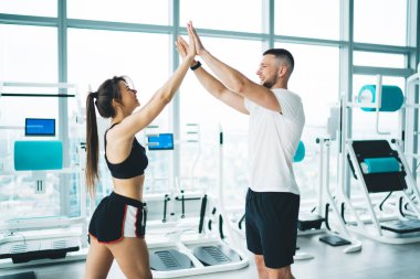 Aktif giyinen sporcu kadının yan görünüşü, gülümseyen partnerin birbirine bakarken spor makinelerine karşı beş puan vermesi.